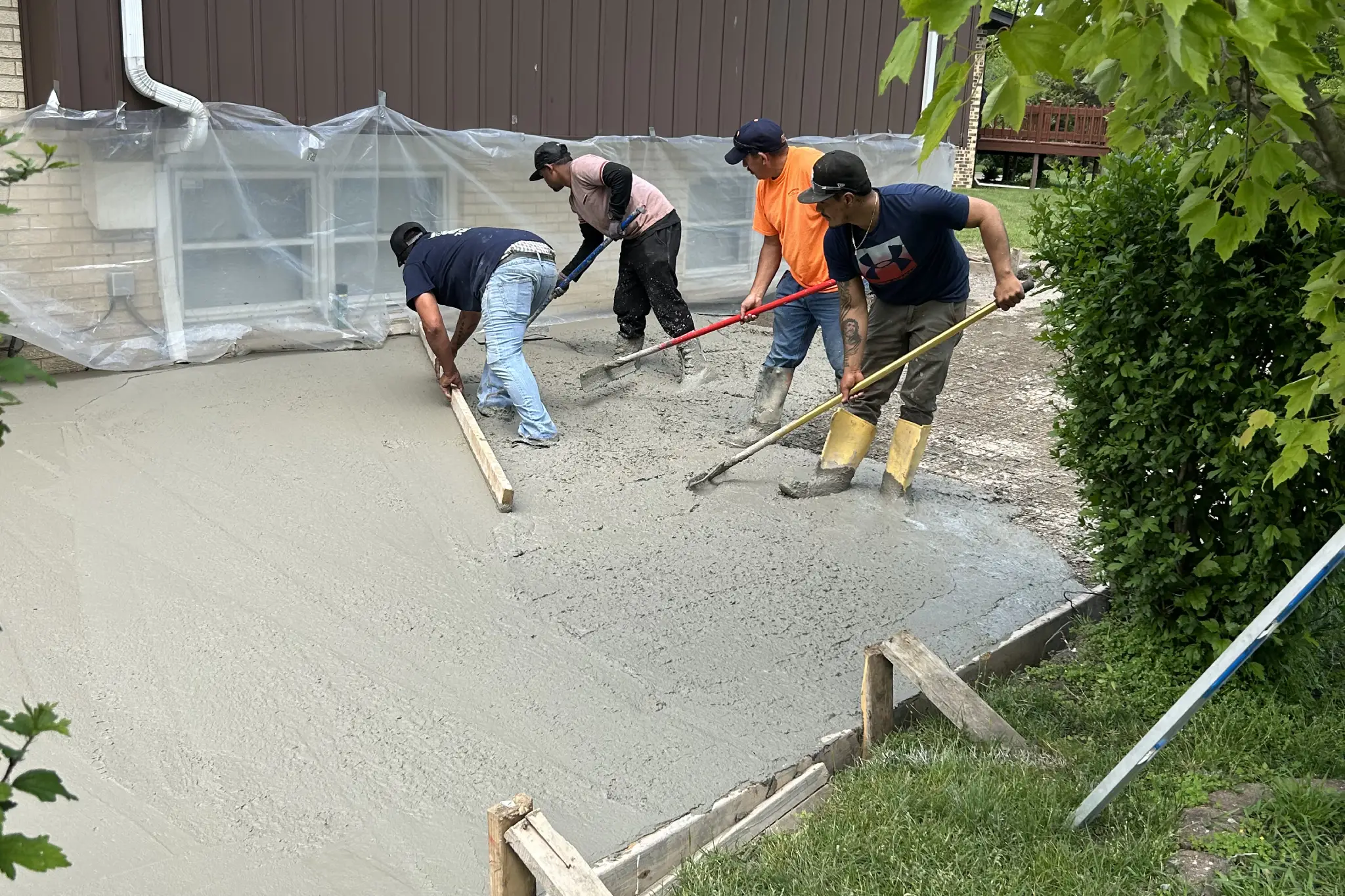 Four workers are smoothing freshly poured concrete on a home site, surrounded by greenery and covered windows