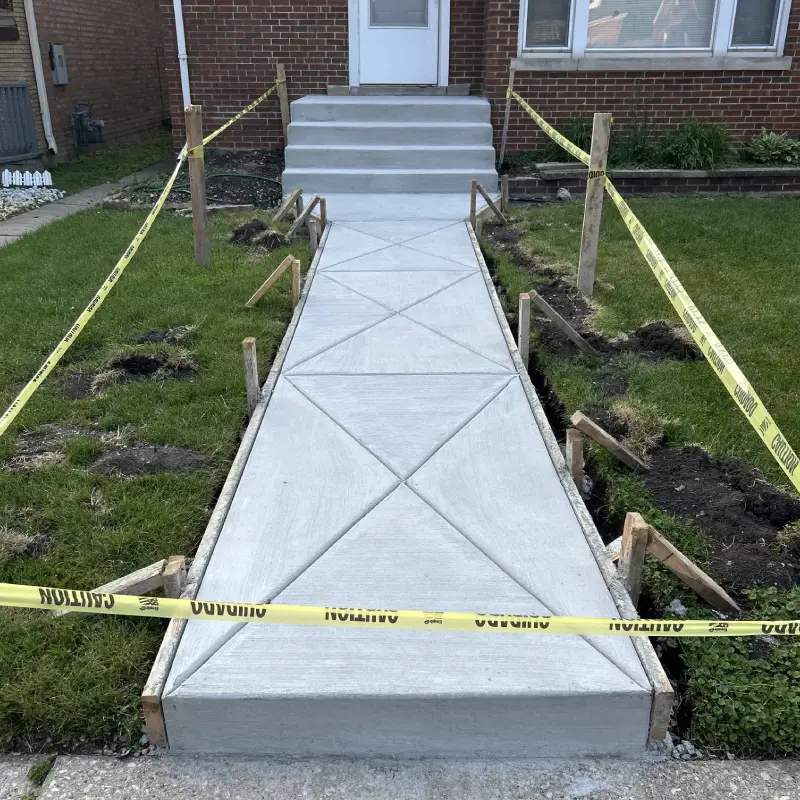 A newly poured concrete walkway leads to a house's entrance, surrounded by caution tape and wooden supports, with freshly cut grass on either side